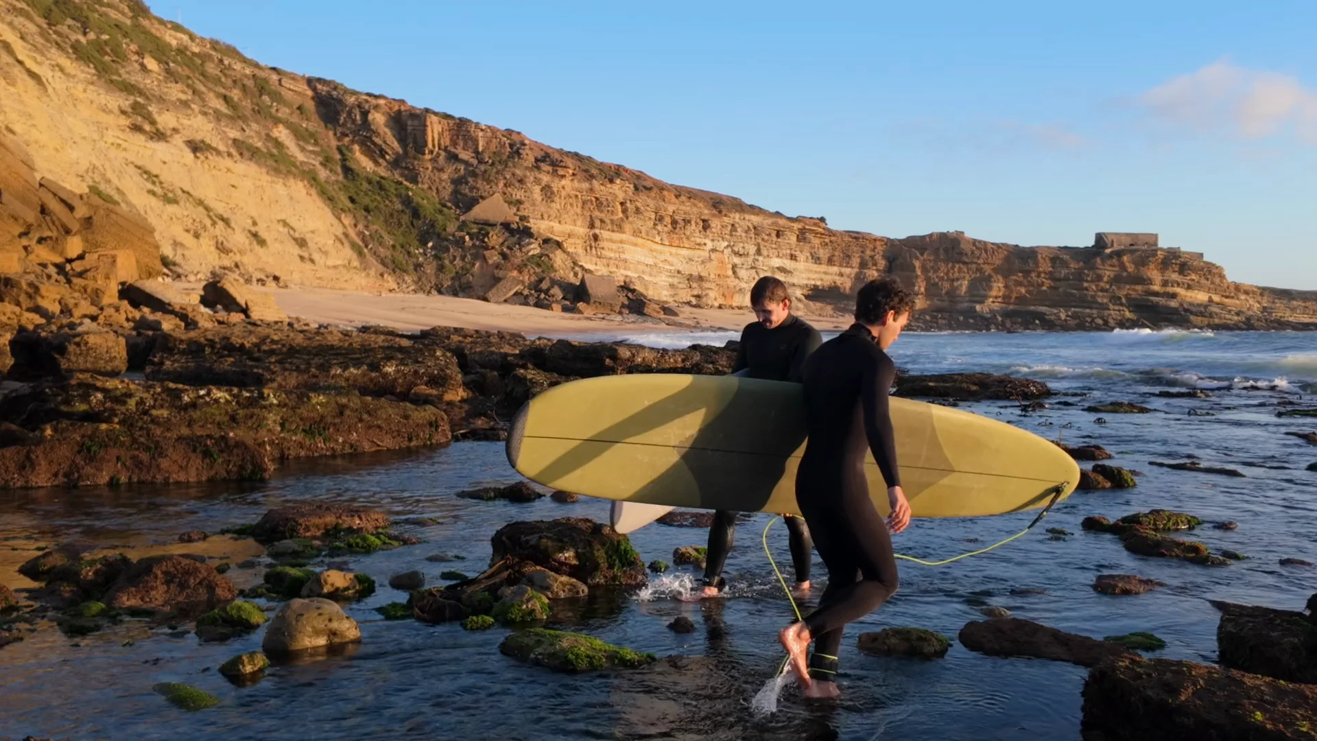 Two surfers carrying their boards into the water beneath golden coastal cliffs.